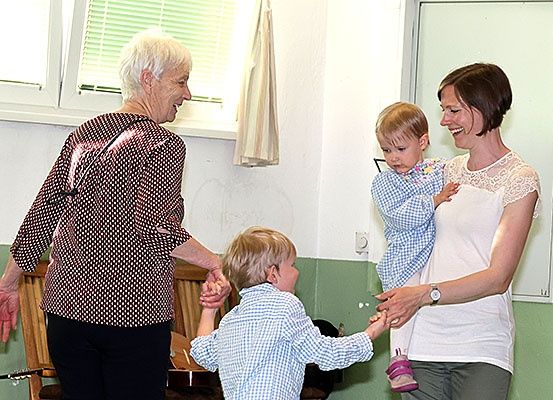 Eine ältere Frau und ein junger Junge halten sich an den Händen und lächeln, in der Nähe einer Frau, die ein Baby hält. Sie sind drinnen, mit einem Fenster mit Jalousien und einer grünen Wand im Hintergrund.