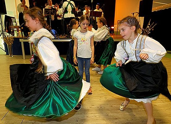 Mehrere Mädchen tanzen auf einem Holzboden, gekleidet in traditionelle Kostüme. Eine Mädchen in der Mitte trägt ein weißes Shirt. Im Hintergrund spielt eine Gruppe von Musikern verschiedene Instrumente auf einer Bühne.