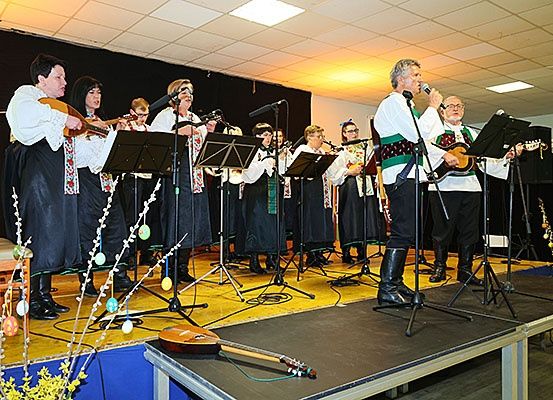 Eine Gruppe von Musikern in traditioneller Kleidung tritt auf einer Bühne auf, einige spielen Saiteninstrumente und andere Blasinstrumente. Die Bühne ist mit Musikinstrumenten und Ostereiern geschmückt.