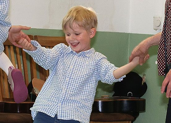 Ein junger Junge mit blonden Haaren, der ein blau kariertes Hemd und blaue Jeans trägt, lächelt und tanzt mit zwei Erwachsenen, einer hält seine linke Hand und der andere seine rechte Hand.