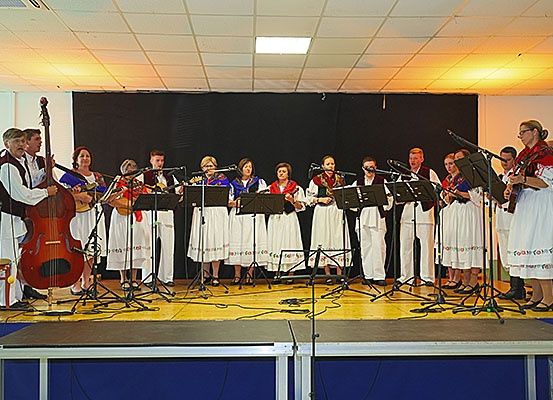 Eine Gruppe von Musikern, einige spielen Saiteninstrumente, andere halten Mikrofone, performen auf einer Bühne mit schwarzem Hintergrund und Deckenbeleuchtung.