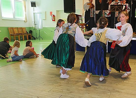 Vier Mädchen in traditionellen Kleidern tanzen auf einem Holzboden, während andere zusehen und eine Band hinter ihnen spielt.
