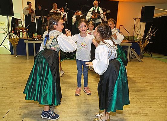 Eine Gruppe von Kindern und Erwachsenen in traditioneller Kleidung tanzt auf einem Holzboden. Ein Mädchen in einem weißen T-Shirt steht zwischen zwei Mädchen in grünen Kleidern. Ein Mann spielt Gitarre hinter ihnen.