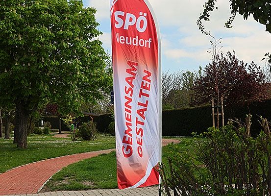 Ein Banner mit den Worten 'Gemeinsam Gestalten' steht auf einem roten Ziegelweg in einem Park mit grünem Rasen und Bäumen.