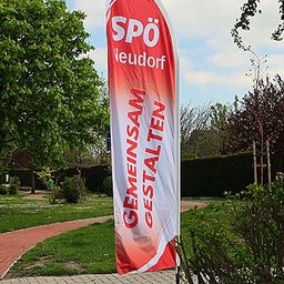 Ein Banner mit den Worten 'Gemeinsam Gestalten' steht auf einem roten Ziegelweg in einem Park mit grünem Rasen und Bäumen.
