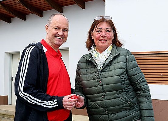 Ein Mann und eine Frau stehen vor einem Gebäude, lächeln und halten ein rotes Objekt. Der Mann trägt ein rotes Shirt und eine schwarze Jacke mit weißen Streifen. Die Frau trägt eine grüne Jacke und hat eine Schürze um den Hals. Hinter ihnen befindet sich eine weiße Wand mit einer Tür und einer braunen Holzwand.