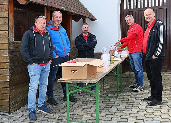 Fünf Männer stehen vor einem Tisch mit Essen und Getränken. Sie tragen alle Jacken. Der Mann auf der linken Seite hat eine Sonnenbrille auf dem Kopf.