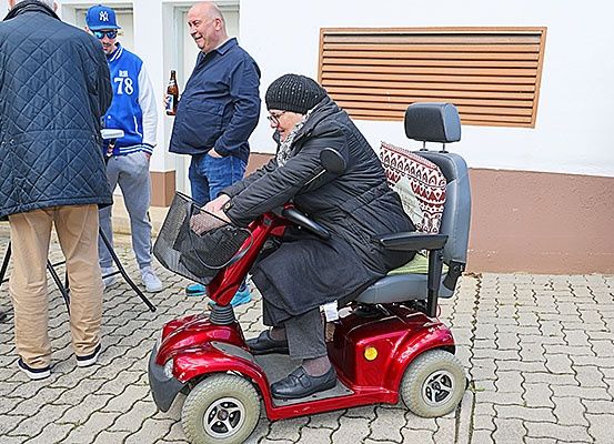 Eine ältere Frau fährt einen roten Roller vor einem Gebäude. Hinter ihr stehen zwei Männer und unterhalten sich, einer hält eine Flasche. Ein anderer Mann, der eine blaue Jacke und eine Sonnenbrille trägt, steht links.