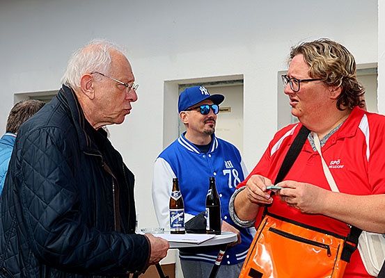 Drei Männer unterhalten sich. Der Mann auf der linken Seite trägt eine Brille und eine dunkle Jacke. Der Mann in der Mitte trägt eine blaue Kappe und eine Sonnenbrille. Der Mann auf der rechten Seite trägt ein rotes Hemd und trägt eine orange Tasche. Auf einem Tisch stehen zwei Flaschen.