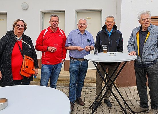 Fünf Personen stehen vor einem Gebäude und lächeln für ein Foto. Drei Männer und zwei Frauen. Sie sind in der Nähe eines Tisches mit Getränken. Hinter ihnen befinden sich zwei Türen.