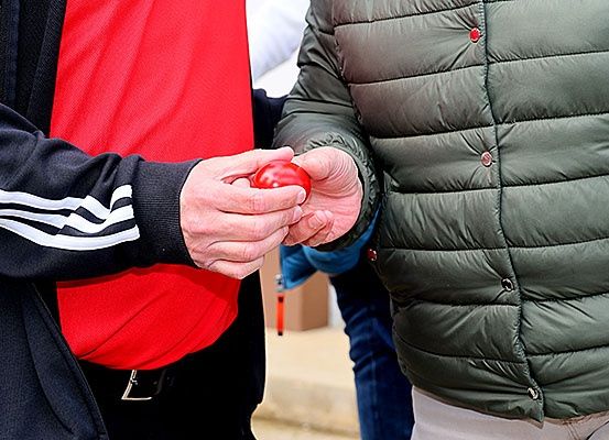 Zwei Personen stehen nah beieinander, eine hält einen roten Ball, die andere trägt eine grüne Jacke mit Knöpfen und einem Gürtel.