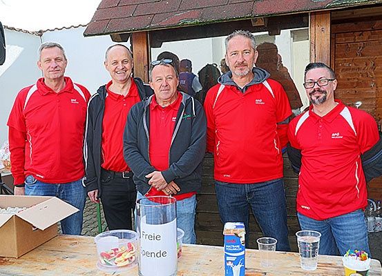Fünf Männer in roten Hemden und blauen Jeans stehen vor einer Holzstruktur. Sie lächeln und scheinen für ein Foto zu posieren. Ein Tisch vor ihnen hat einen Glasbehälter mit dem Wort 'Freie' und verschiedene Gegenstände darauf.