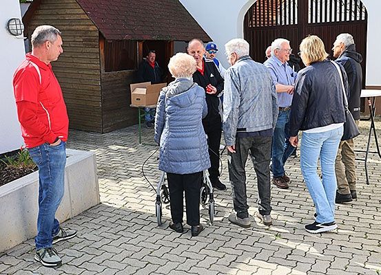 Mehrere Menschen stehen in einem Innenhof, darunter eine ältere Frau mit einem Rollator, und unterhalten sich in der Nähe eines kleinen Gebäudes und eines Tisches.