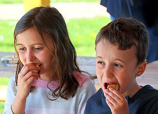 Zwei Kinder essen draußen an einer Picknickbank, eines scheint verärgert, während das andere ruhig isst.