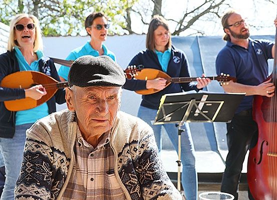Eine Gruppe von Musikern spielt im Freien. Vier Personen spielen Saiteninstrumente, darunter ein Cello, während ein Mann in einer Baskenmütze zuschaut.