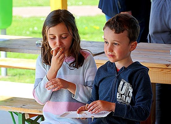 Zwei Kinder sitzen an einer Picknickbank und essen. Das Mädchen trägt einen Pullover, der Junge eine Kapuzenjacke. Hinter ihnen stehen zwei Erwachsene an einem Tisch.