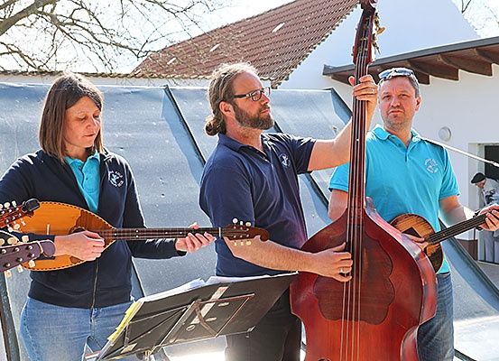 Eine Gruppe von Menschen spielt verschiedene Saiteninstrumente, darunter eine Mandoline, eine Gitarre und einen Bass. Sie stehen vor einem Gebäude mit Dach.