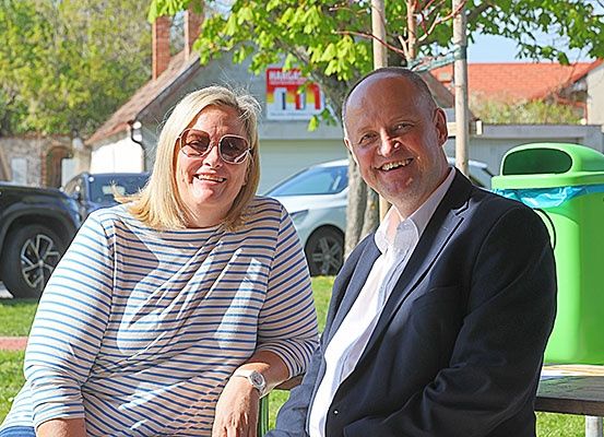 Ein Mann und eine Frau sitzen lächelnd auf einer Parkbank. Hinter ihnen befinden sich Häuser, Autos und ein grüner Mülleimer.