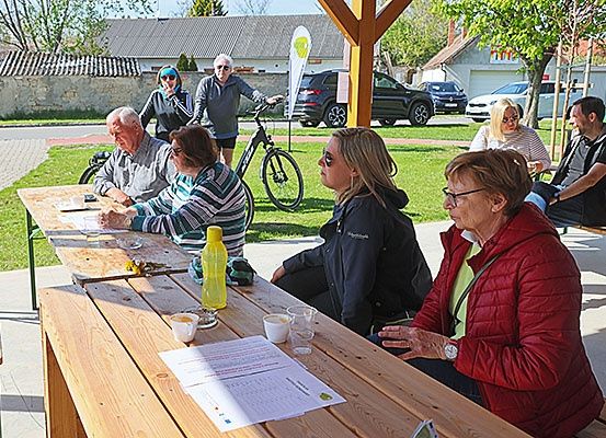 Eine Gruppe von Menschen sitzt um einen Holztisch in einem Park. Ein Mann steht neben einem Fahrrad. Papiere und Tassen sind auf dem Tisch.