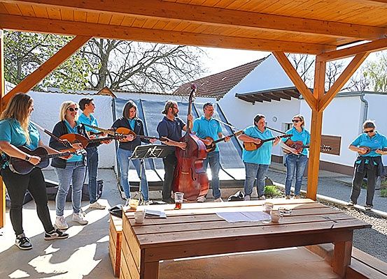 Eine Gruppe von Menschen in blauen Shirts spielt verschiedene Saiteninstrumente auf einer Terrasse unter einer Holzstruktur. Die Instrumente umfassen Gitarren, einen Cello und eine Mandoline. Ein Tisch mit Gläsern steht in der Nähe. Die Umgebung ist draußen mit Bäumen und Gebäuden im Hintergrund.