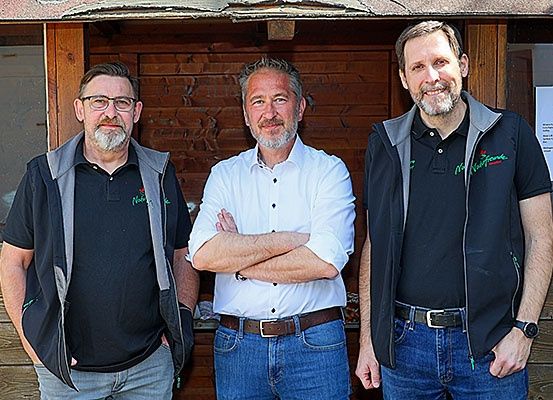 Drei Männer stehen lächelnd vor einer Holzstruktur. Der Mann auf der linken Seite trägt eine Brille und eine Jacke, während der Mann in der Mitte ein weißes Hemd und verschränkte Arme hat. Der Mann auf der rechten Seite trägt ein schwarzes Shirt mit Logo und einer Uhr.
