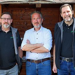 Drei Männer stehen lächelnd vor einer Holzstruktur. Der Mann auf der linken Seite trägt eine Brille und eine Jacke, während der Mann in der Mitte ein weißes Hemd und verschränkte Arme hat. Der Mann auf der rechten Seite trägt ein schwarzes Shirt mit Logo und einer Uhr.