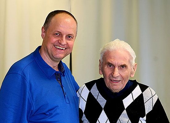 Zwei Männer stehen nebeneinander und lächeln für ein Foto. Der Mann auf der linken Seite trägt ein blaues Poloshirt. Der Mann auf der rechten Seite trägt einen schwarzen und weißen Rautenpullover. Sie schauen beide in die Kamera. Hinter ihnen ist ein weißer Vorhang.