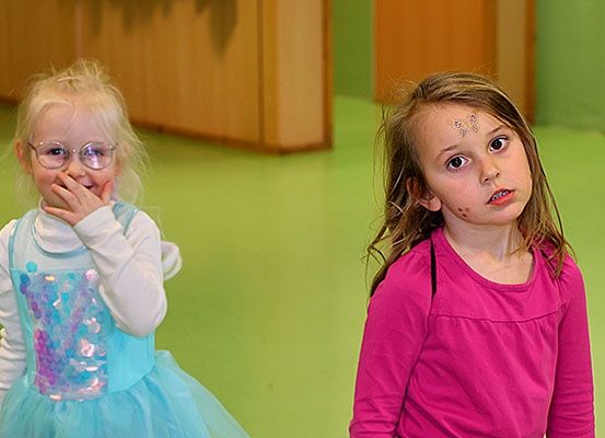 Zwei junge Mädchen stehen nebeneinander; eine trägt eine Brille und ein blaues Kleid, die andere ein rosa Hemd mit Schmetterlings-Gesichtsbemalung.