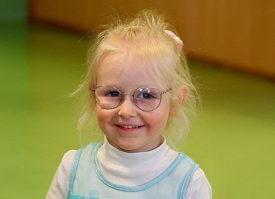 Ein kleines Mädchen mit blonden Haaren und Brille lächelt in die Kamera. Sie trägt ein weißes Rollkragenshirt und ein blaues Kleid. Der Hintergrund ist grün.