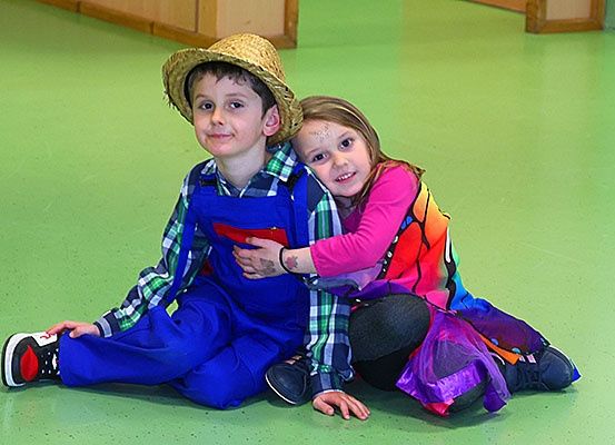 Zwei Kinder sitzen auf einem grünen Boden und lächeln, wobei der Junge einen Strohhut trägt und das Mädchen ein rosa Hemd trägt.