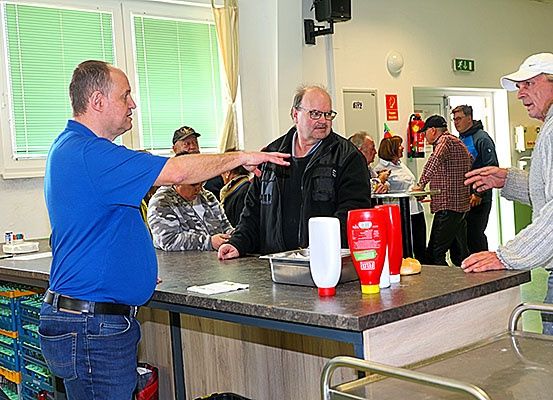 Drei Männer stehen an einer Theke in einer Cafeteria. Ein Mann in einem blauen Shirt zeigt, während ein anderer Mann in einem schwarzen Mantel zusieht. Ein Mann in einer weißen Kappe lächelt.