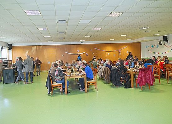 Eine Gruppe von Menschen sitzt an Tischen in einem Essbereich mit Holzwänden und grünem Boden. Einige Leute stehen in der Nähe der Wand und einige gehen. An der Wand befinden sich Lautsprecher und Dekorationen.