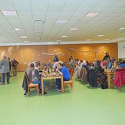 Eine Gruppe von Menschen sitzt an Tischen in einem Essbereich mit Holzwänden und grünem Boden. Einige Leute stehen in der Nähe der Wand und einige gehen. An der Wand befinden sich Lautsprecher und Dekorationen.