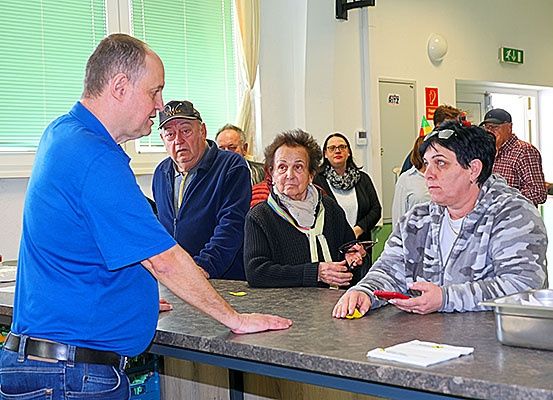 Ein Mann in einem blauen Shirt steht hinter einem Tresen und spricht mit einer Frau in einer grauen Kapuzenjacke. In der Nähe unterhalten sich mehrere Menschen, darunter eine ältere Frau in einem schwarzen Pullover und ein Mann mit einer Mütze.