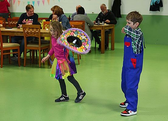 Ein junges Mädchen und ein Junge spielen mit einem bunten Hut bzw. einer Augenmaske in einem Raum mit grünem Boden. Hinter ihnen sitzen Menschen an Tischen.