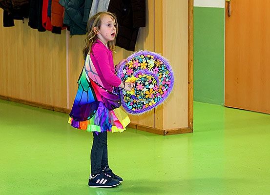 Ein kleines Mädchen in einem Schmetterlingskostüm steht in einem Raum, hält eine bunte Windmühle und schaut nach oben. Hinter ihr hängen Kleidungsstücke an einer Wand und zwei Holztüren sind sichtbar.