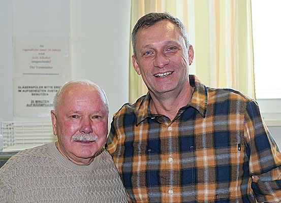 Zwei Männer stehen eng beieinander und lächeln für ein Foto. Sie stehen in einem Raum mit einem Fenster und Vorhängen hinter ihnen.