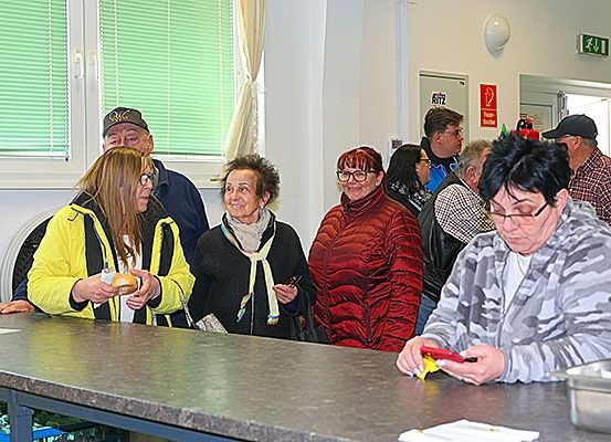 Mehrere Menschen, darunter ältere Personen, stehen um einen Tresen in einem Raum. Eine Person hält Essen, und eine andere benutzt ein Handy. Es gibt Fenster und ein Feuerlöscher-Schild an der Wand.