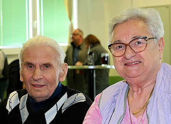 Zwei ältere Personen, ein älterer Mann und eine ältere Frau, sitzen zusammen, lächeln und posieren für ein Foto. Im Hintergrund, unscharf, sind Menschen zu sehen, die in der Nähe eines Tisches mit einer Flasche stehen.