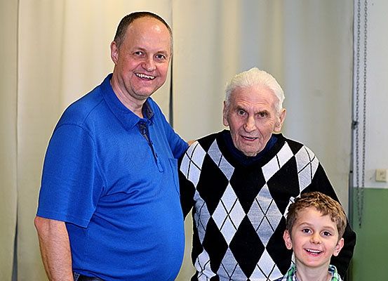 Ein Mann in einem blauen T-Shirt steht zwischen einem älteren Mann und einem jungen Jungen, alle drei lächeln und posieren für ein Foto in einem Raum mit einem weißen Vorhang.