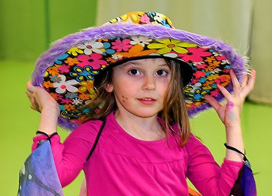 Ein kleines Mädchen trägt einen bunten Blumenhut und ein rosa Shirt und hält den Hut mit beiden Händen fest.