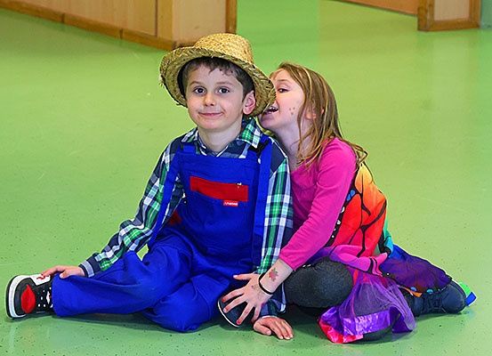 Zwei Kinder sitzen auf einem grünen Boden, ein Junge trägt einen Strohhut und ein blaues Outfit, und ein Mädchen in einem rosa Langarmshirt und einem farbenfrohen Kleid. Sie lächeln beide und scheinen sich zu amüsieren.
