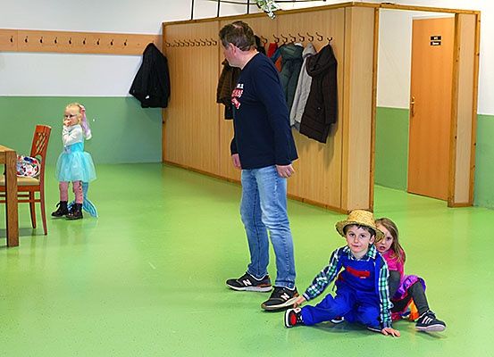 Ein Mann steht zwischen zwei sitzenden Kindern, einem Mädchen und einem Jungen, während ein Mädchen hinter ihm steht. Sie befinden sich in einem Raum mit grünem Boden und Wänden, Holzschränken und hängenden Mänteln.