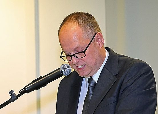 Ein Mann in Anzug und Brille spricht in ein Mikrofon in einem Konferenzraum.