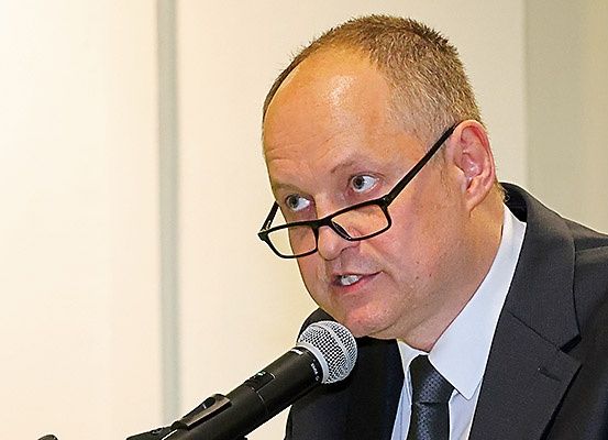 Ein Mann in einem schwarzen Anzug und mit Brille spricht in ein Mikrofon.