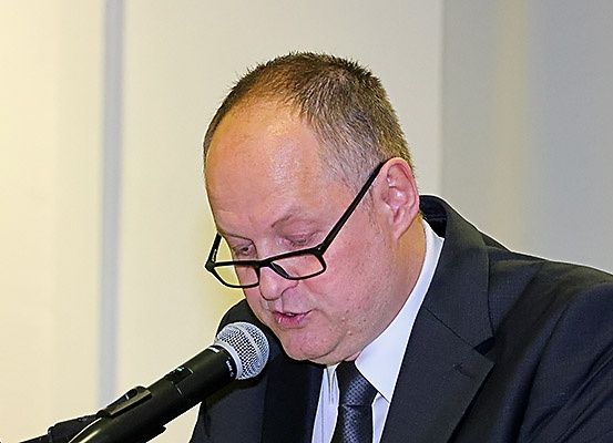 Ein Mann in einem schwarzen Anzug und mit Brille spricht in ein Mikrofon, wahrscheinlich auf einer Konferenz. Er hat einen ernsten Ausdruck.