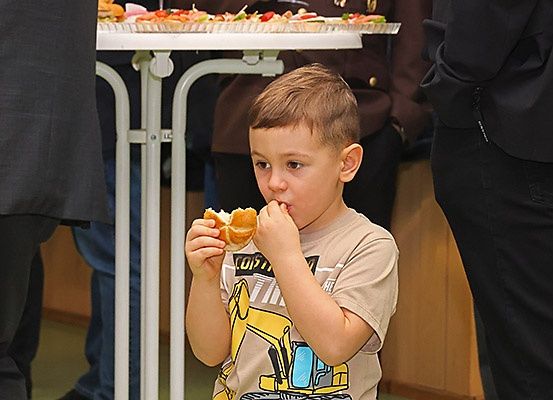 Ein kleiner Junge isst ein Sandwich vor einem Buffet-Tisch. Er trägt ein beiges T-Shirt mit einem gelben Baufahrzeug-Design.