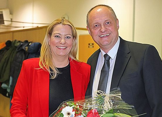 Ein Mann und eine Frau stehen eng beieinander und lächeln. Der Mann hält einen Blumenstrauß. Sie sind formell gekleidet, und die Frau trägt einen roten Blazer. Hinter ihnen hängen Kleidungsstücke an einem Kleiderständer.