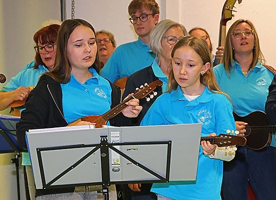 Eine Gruppe von Musikern in blauen Hemden spielt verschiedene Instrumente in einem Raum. Zwei Mädchen vorne spielen Gitarre.