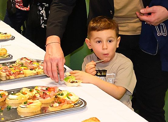 Ein kleiner Junge isst ein Sandwich an einem Buffettisch. Auf dem Tisch sind verschiedene Speisen, und Erwachsene stehen herum.